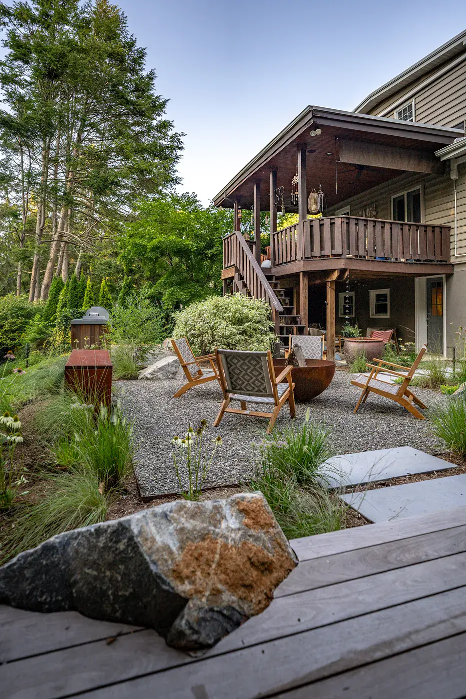 Twilight view from the wood deck across a Havertown backyard landscape design, with a boulder in the foreground, a steel fire pit and lounge chairs on a gravel patio, bluestone steppers, ornamental grasses, and an outdoor kitchen below the existing covered deck