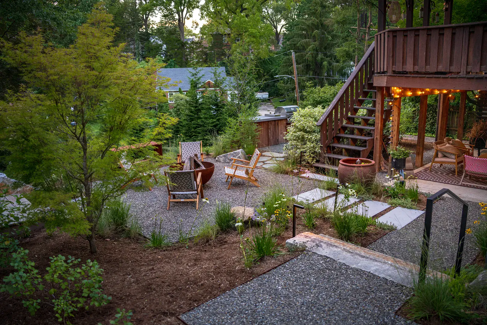 Elevated dusk view of a Havertown backyard landscape design with a steel fire pit and lounge chairs on a gravel patio, bluestone stepping stones, a granite step with black steel railing, a Japanese maple, and an outdoor kitchen beside the existing deck