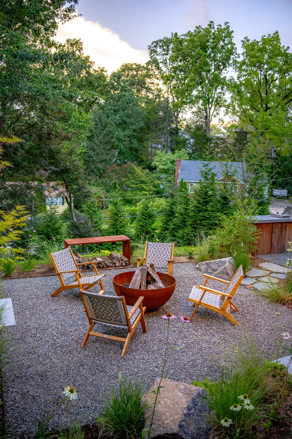 Steel fire pit with stacked firewood and four lounge chairs on a gravel patio in a Havertown backyard landscape design, with boulders, echinacea, ornamental grasses, a steel log bench, and an outdoor kitchen at dusk