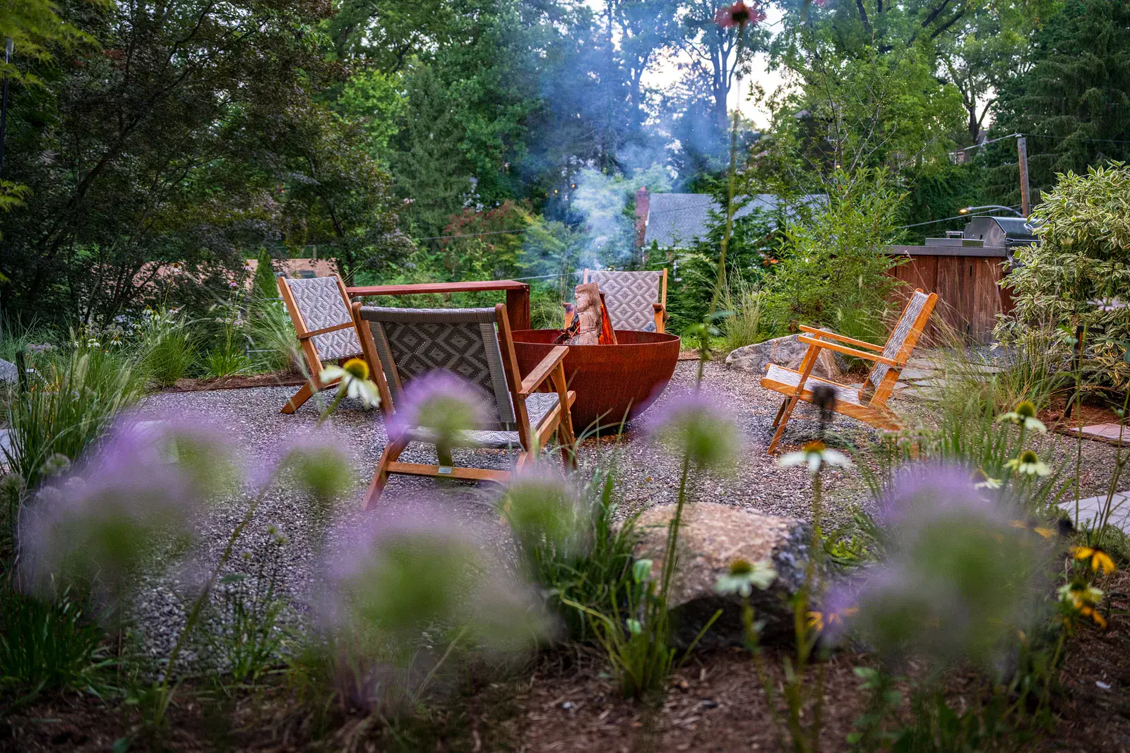 Weathered steel fire bowl with smoke rising, surrounded by woven teak lounge chairs on a gravel patio, with boulders, allium, echinacea, and ornamental grasses in the foreground and an outdoor kitchen beyond