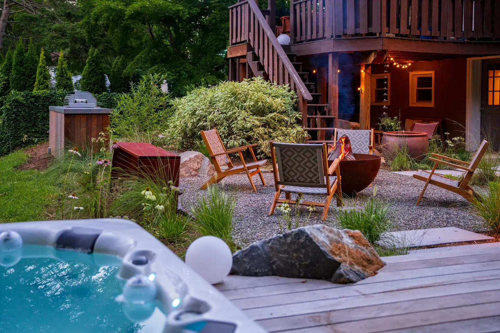 Dusk view from the hot tub and wood deck across a Havertown backyard landscape design, with a lit steel fire pit and lounge chairs on a gravel patio, ornamental grasses, a boulder, an outdoor kitchen, and string lights under the existing deck
