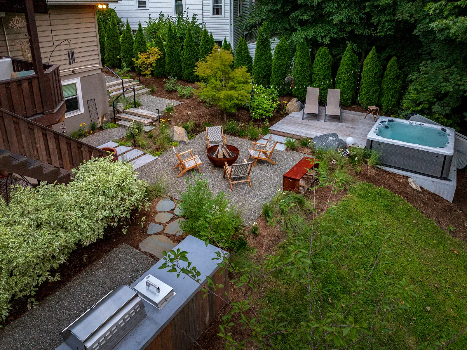 Aerial dusk view of a Havertown backyard landscape design with a steel fire pit and lounge chairs on a gravel patio, a hot tub and chaise lounges on a wood deck, granite steps with black steel railings, an outdoor kitchen, and a screen of evergreens