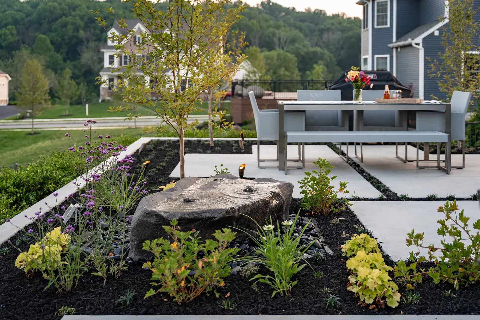 Bubbling boulder water feature on black river stone with purple verbena, chartreuse heuchera, and a young tree beside concrete walkways leading to a stainless and gray outdoor dining table and bench in a Newtown Square PA landscape design