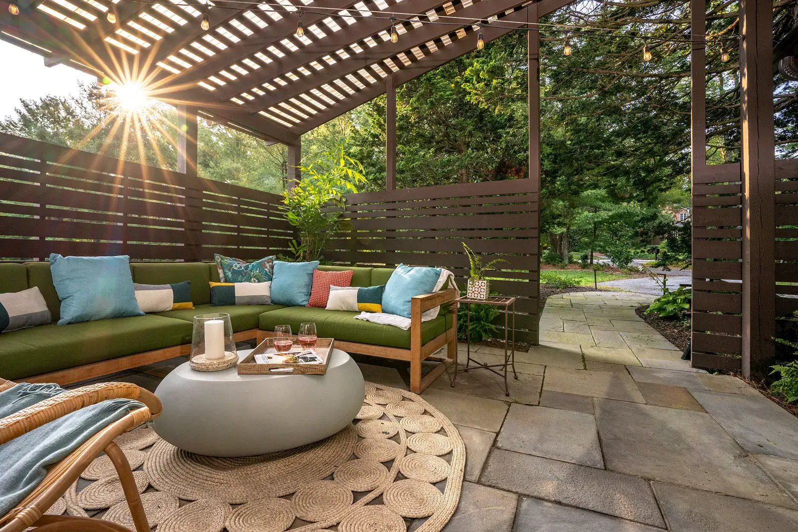 Sunlit view from a pergola patio with a green sectional and rope rug through horizontal slat screening to a bluestone path leading to the driveway in a Wilmington DE private garden design
