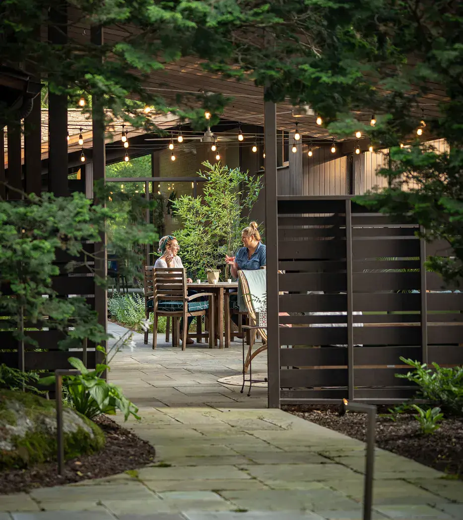 Two women dining at a teak table on a lit bluestone patio, seen through slat screening, a bluestone path, and evergreen branches in a Wilmington DE private garden design