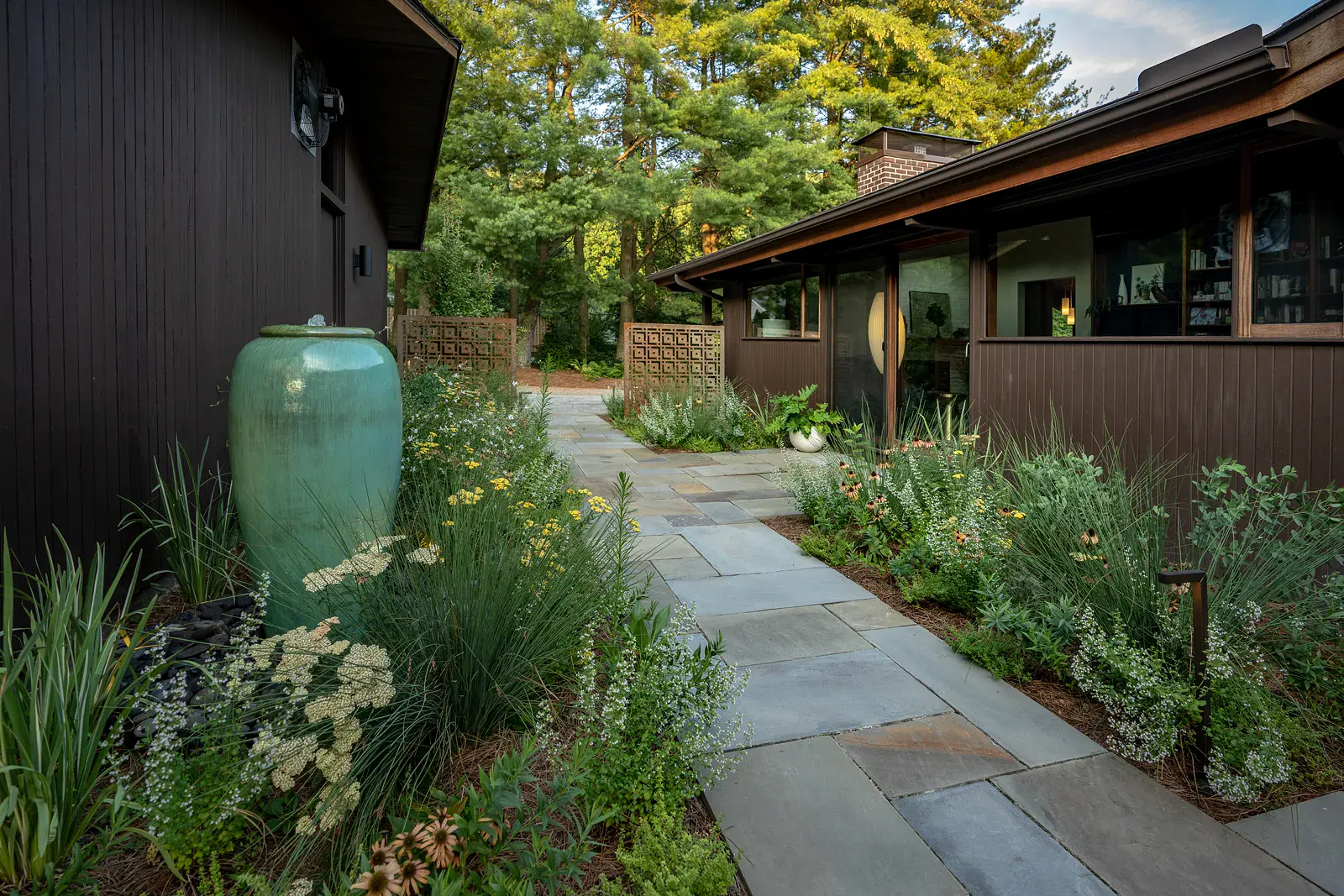 Bluestone breezeway walkway lined with ornamental grasses, yarrow, echinacea, and calamint, a glazed ceramic fountain urn, and Corten steel screens beside a mid-century modern home in Wilmington DE