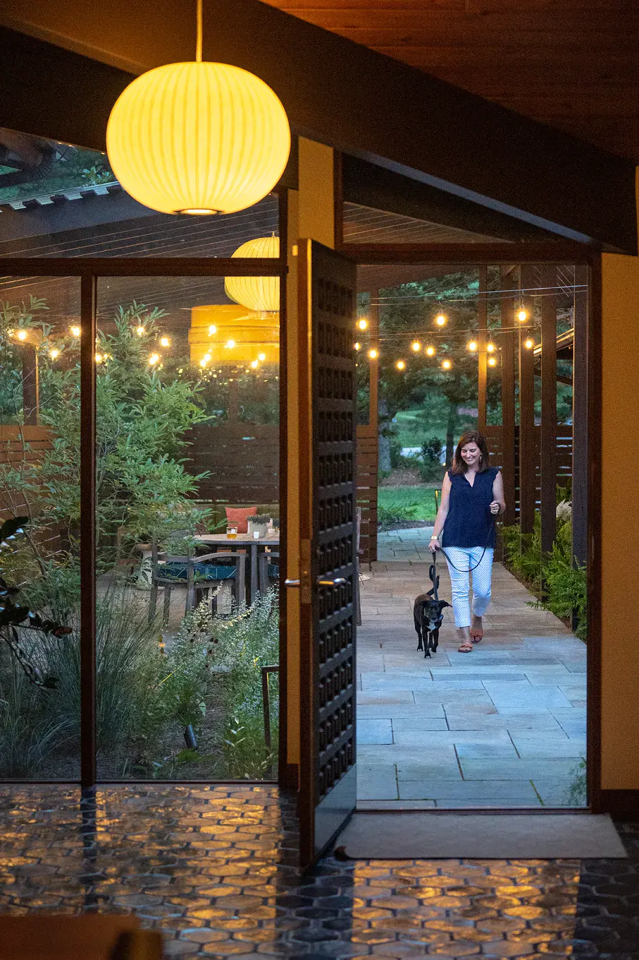 Woman walking her dog along a bluestone breezeway with string lights, seen through an open paneled door with a glowing globe pendant, toward the garden patio in a Wilmington DE landscape design