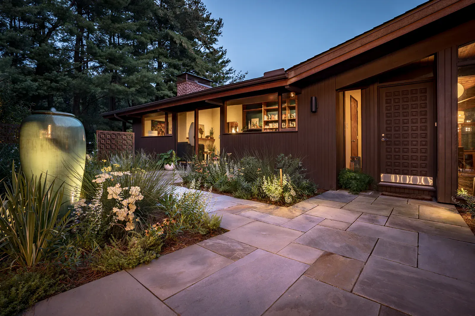 Dusk view of a Wilmington DE entry garden with an uplit ceramic fountain urn, Corten steel screen, yarrow and calamint plantings, a bluestone walk, and a paneled wood front door