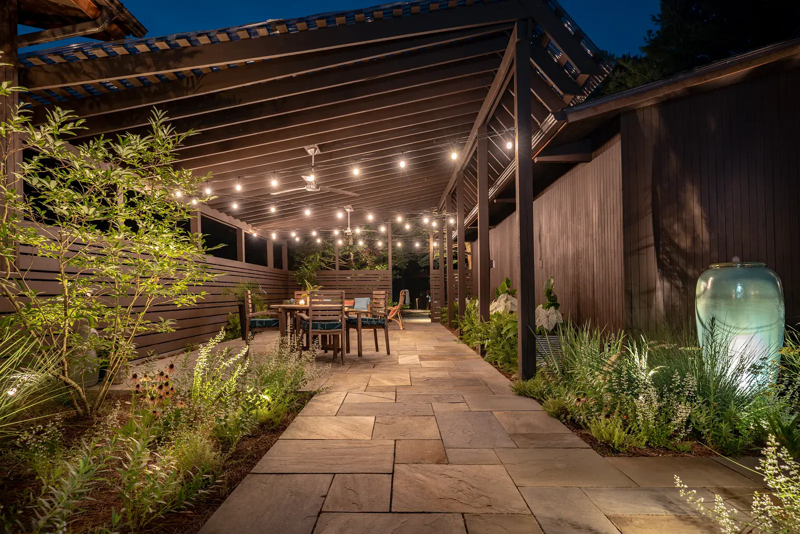 Night view of a bluestone breezeway lined with perennials and an uplit ceramic fountain urn, leading to a string-lit pergola patio with a teak dining table and ceiling fans in a Wilmington DE landscape design