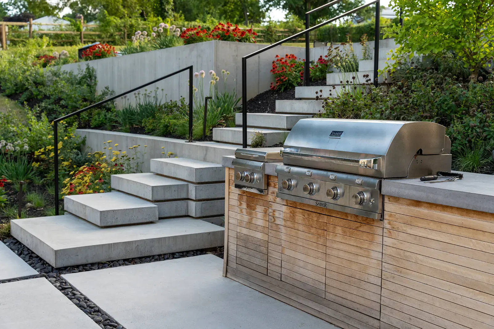 fpo-800x600 Custom outdoor kitchen with a built-in gas grill and wood-slat cabinetry beside offset floating concrete steps and black metal railings in a Newark DE family backyard landscape design, with river stone joints and echinacea, allium, and yarrow plantings