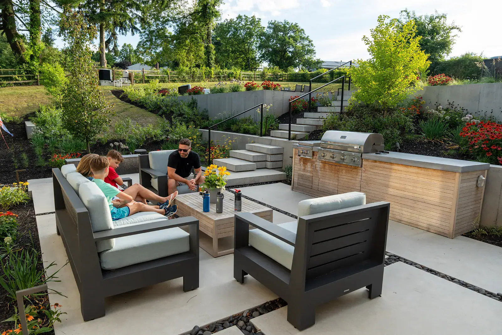 fpo-800x600 A family relaxing on the lower patio of a Newark DE family backyard landscape design, with modern outdoor seating on concrete pavers set in river stone, a custom outdoor kitchen, floating concrete steps with black metal railings, and tiered concrete walls with echinacea and allium plantings