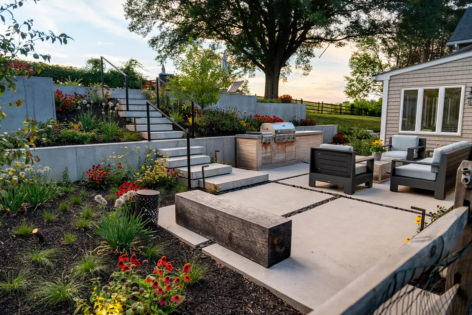Dusk view of a Newark DE family backyard landscape design with offset floating concrete steps and black metal railings rising beside a custom outdoor kitchen, a weathered timber bench, outdoor seating on concrete pavers, and echinacea and allium plantings under a mature oak