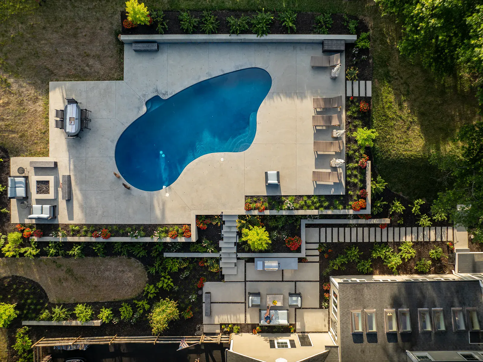 fpo-800x600 Overhead view of a Newark DE family backyard landscape design with a freeform pool, concrete terrace, fire pit lounge, dining and lounger areas, floating concrete steps, outdoor kitchen, and tiered perennial beds stepping down toward the home