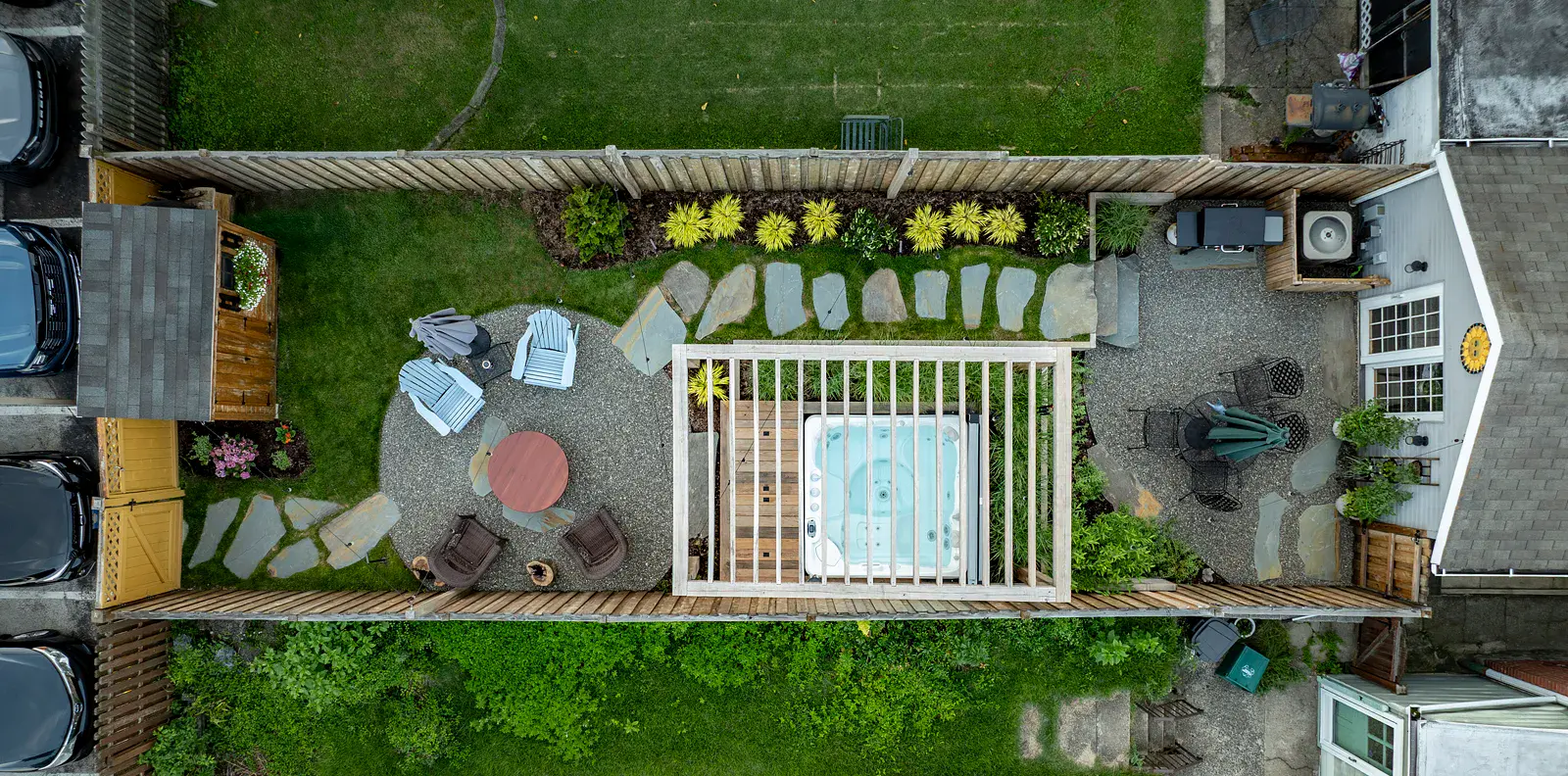 Aerial view of a narrow urban backyard with a pergola-covered hot tub, circular gravel seating area, dining patio, and natural stone paths connecting lawn areas and planted borders within wood privacy fencing.