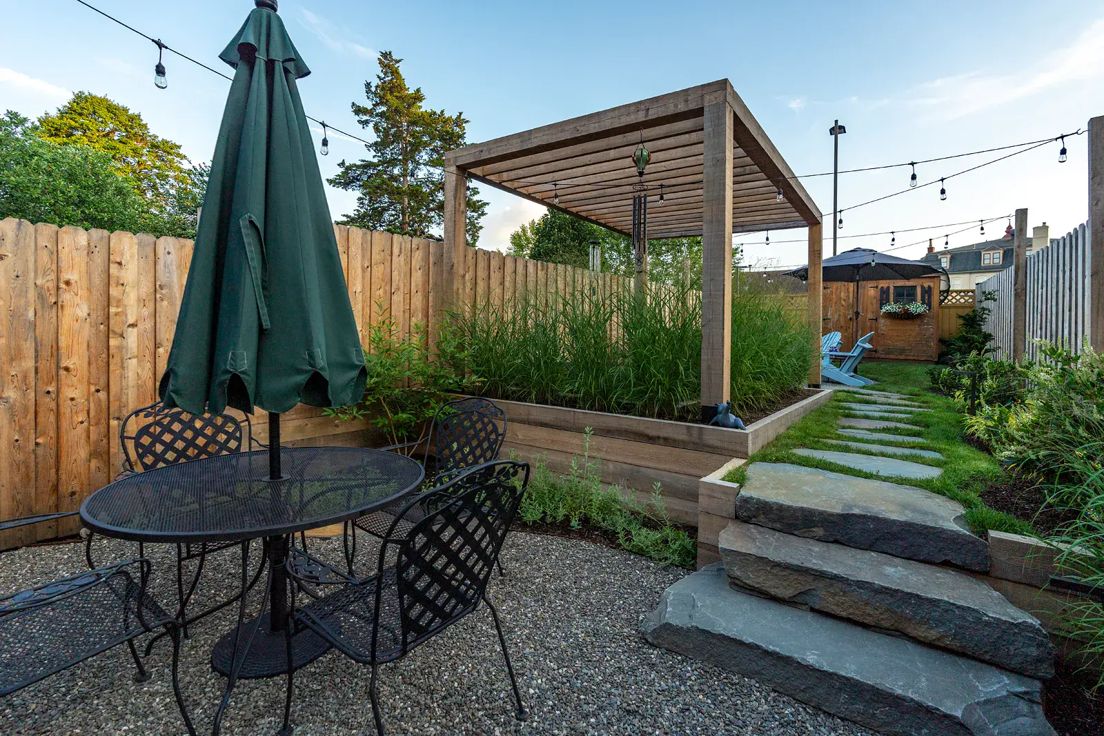 Urban backyard with a gravel dining patio, natural stone steps, and a stepping-stone lawn path beside raised beds of ornamental grasses, a wooden pergola, privacy fencing, and overhead string lights.