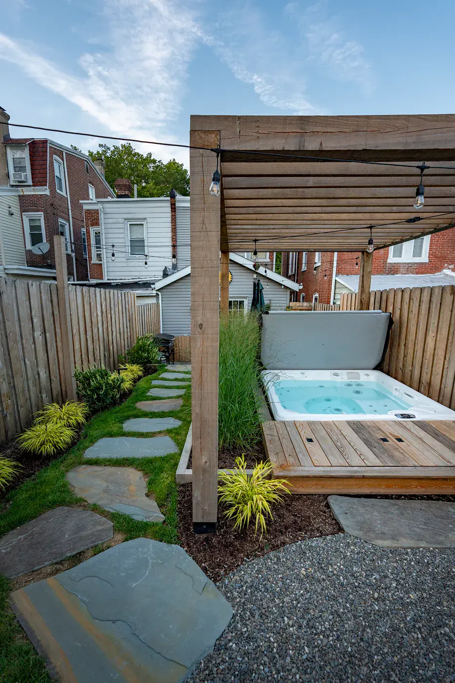 Hot tub beneath a wooden pergola with string lights in a narrow urban backyard, bordered by ornamental grasses, a wood platform, and a natural stone stepping-stone path set into the lawn.