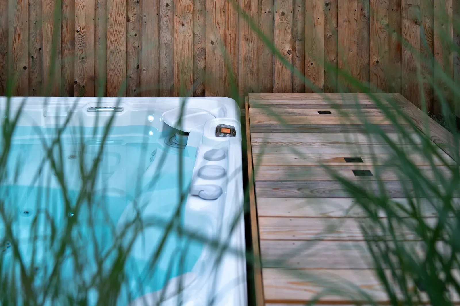 Backyard hot tub with illuminated blue water beside a wooden platform with inset access panels, framed by ornamental grasses and a wood privacy fence.