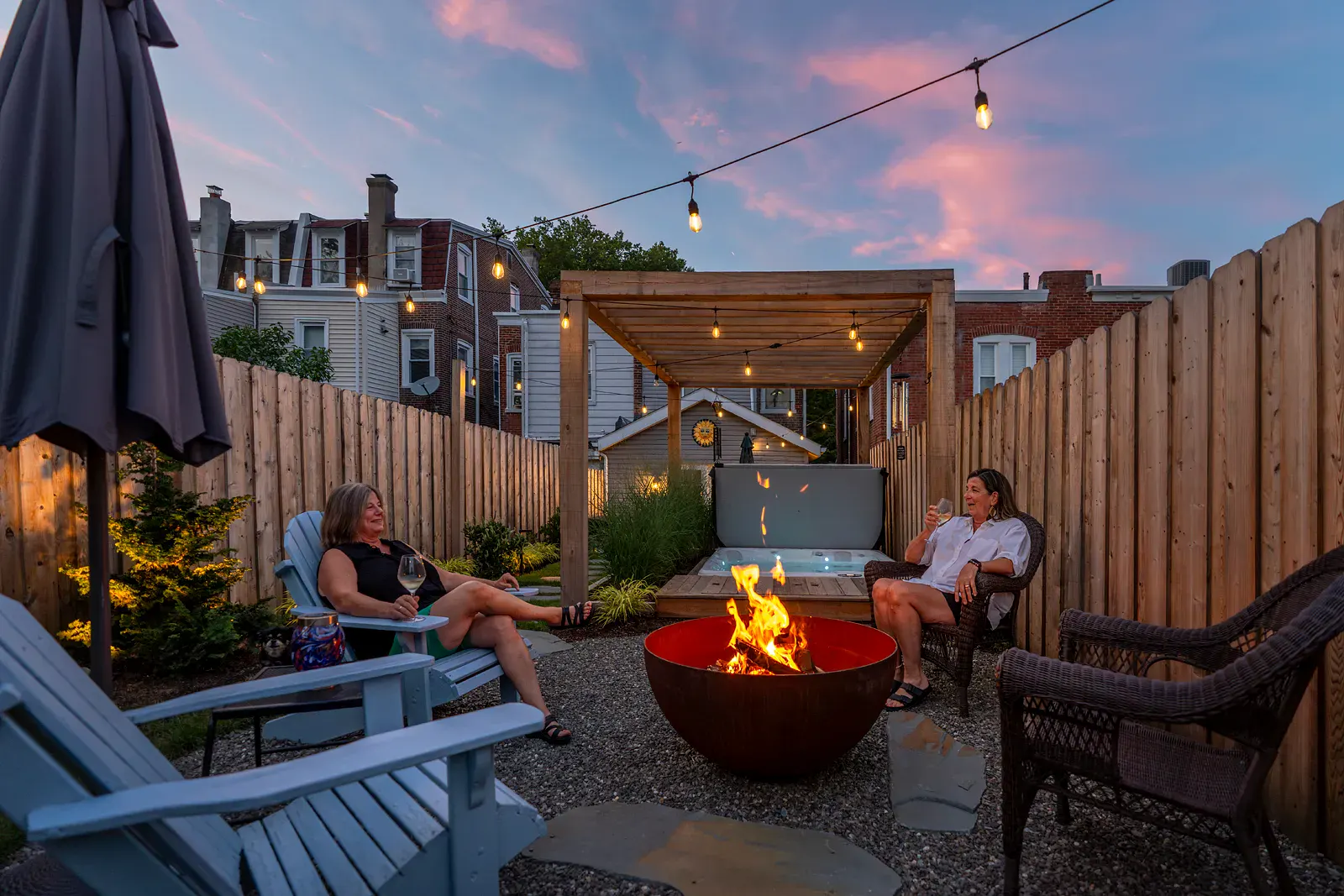 Urban backyard landscape with a gravel fire pit patio, natural stone stepping stones, cedar privacy fencing, and a hot tub beneath a wooden pergola, with two people relaxing under string lights at sunset.