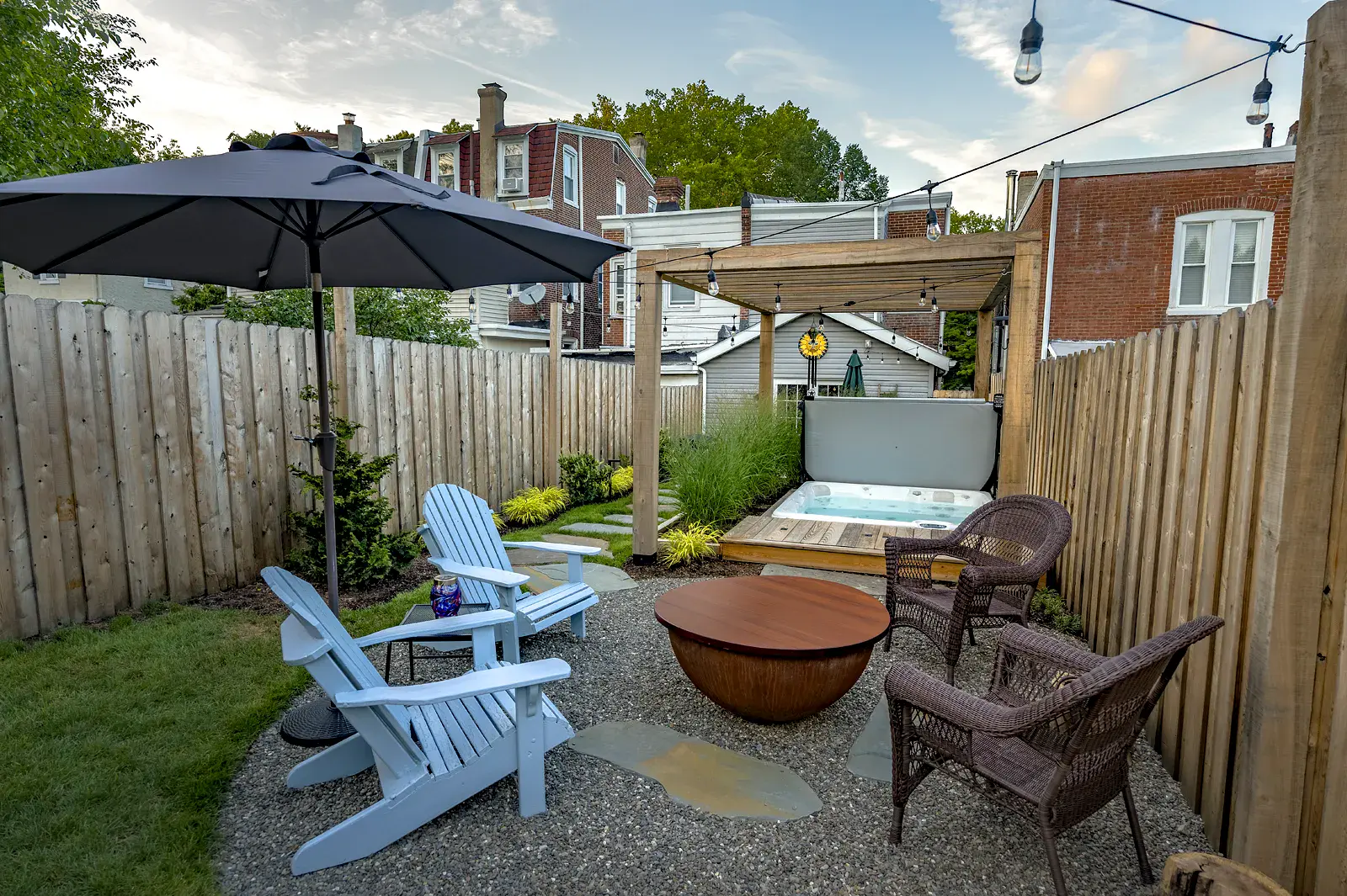Urban backyard gravel patio with blue Adirondack chairs and wicker seating around a covered fire bowl, a large umbrella, and string lights, leading to a pergola-covered hot tub bordered by ornamental grasses and wood privacy fencing.