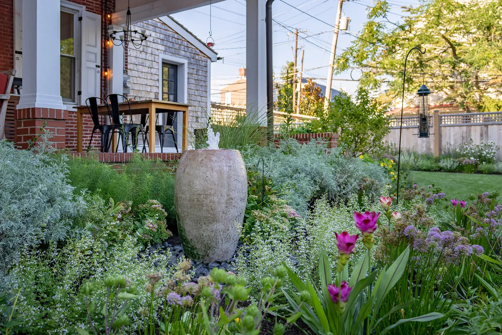 Bubbling ceramic fountain urn on black river stone in a West Chester PA garden design, surrounded by silvery artemisia, pink curcuma, allium, and white calamint, with a covered brick porch, bird feeder, and cedar fence beyond