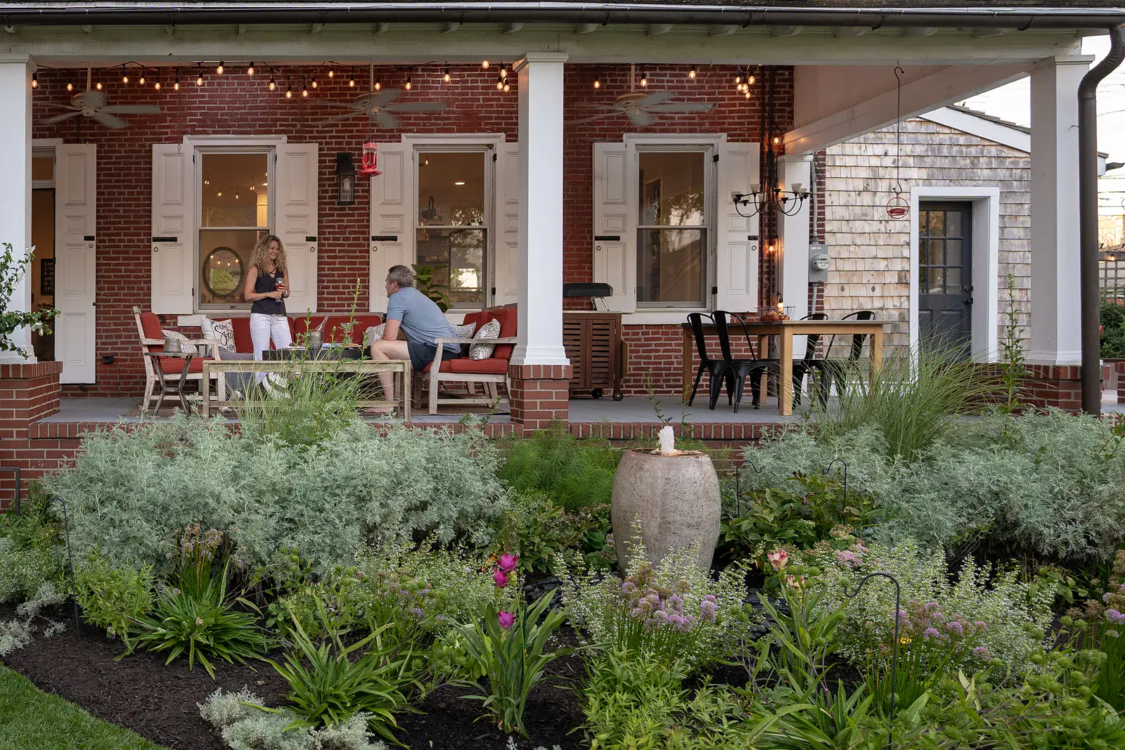 Evening view of a covered brick porch with string lights, ceiling fans, and generous seating in a West Chester PA garden design, with a bubbling fountain urn, silvery artemisia, allium, and pink curcuma in the foreground