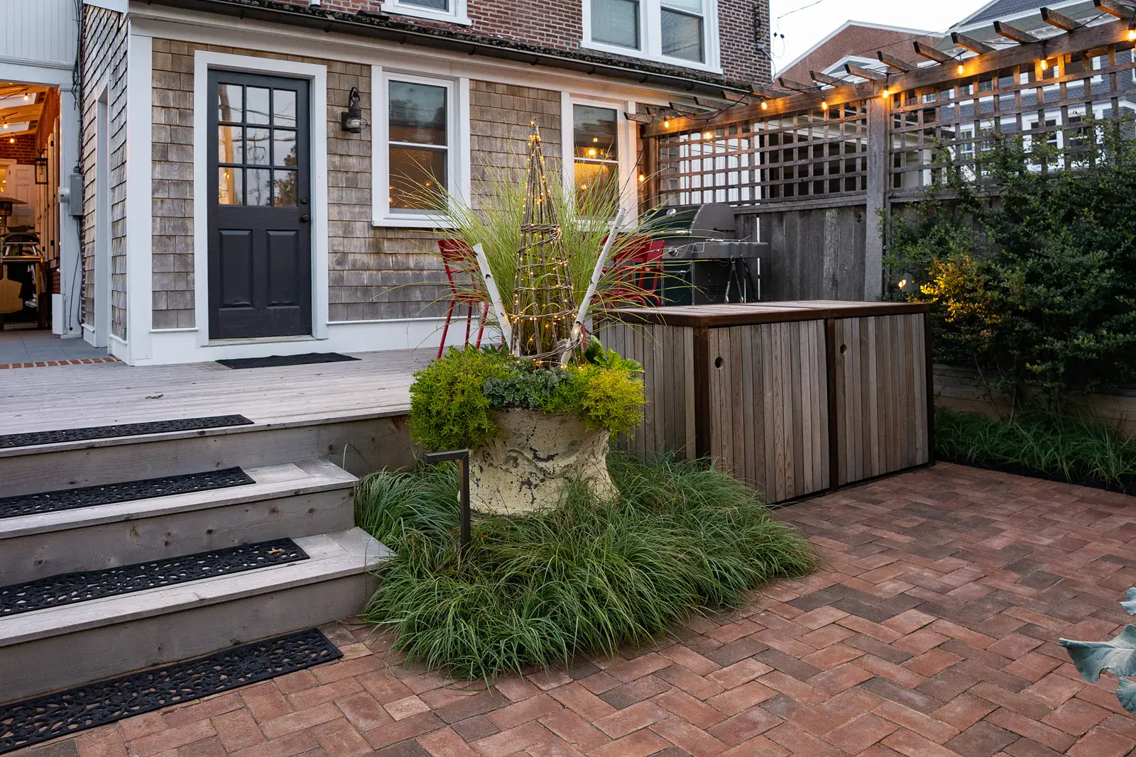 Herringbone brick paver patio beside a wood deck with steps in a West Chester PA garden design, with a large planted urn and string-lit trellis, a custom wood and steel storage cabinet, a timber pergola with string lights, and ornamental grasses