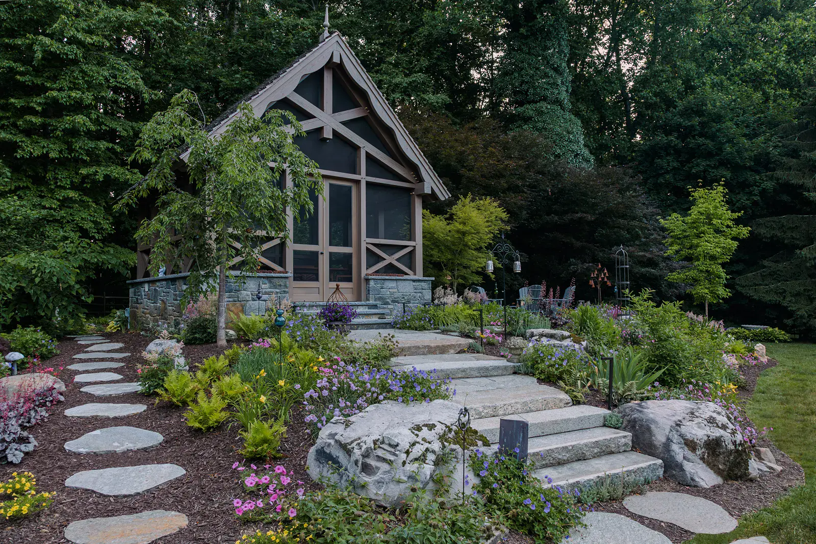 Timber-frame screen room with a stone base, X-bracing, and a gabled cedar shake roof in a Lincoln University PA woodland garden design, reached by irregular stepping stones and wide granite steps between boulders, with ferns, hardy geranium, daylilies, and astilbe