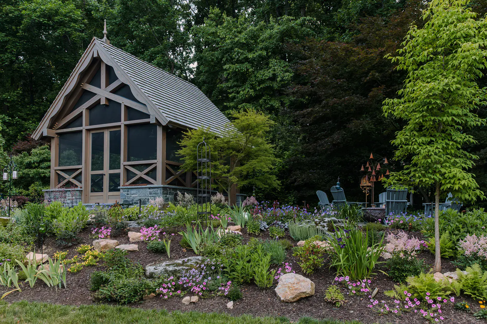 Layered woodland perennial garden of ferns, astilbe, iris, hardy geranium, and petunias among boulders in a Lincoln University PA garden design, rising to a timber-frame screen room with a stone base and a fire pit terrace with gray Adirondack chairs beyond