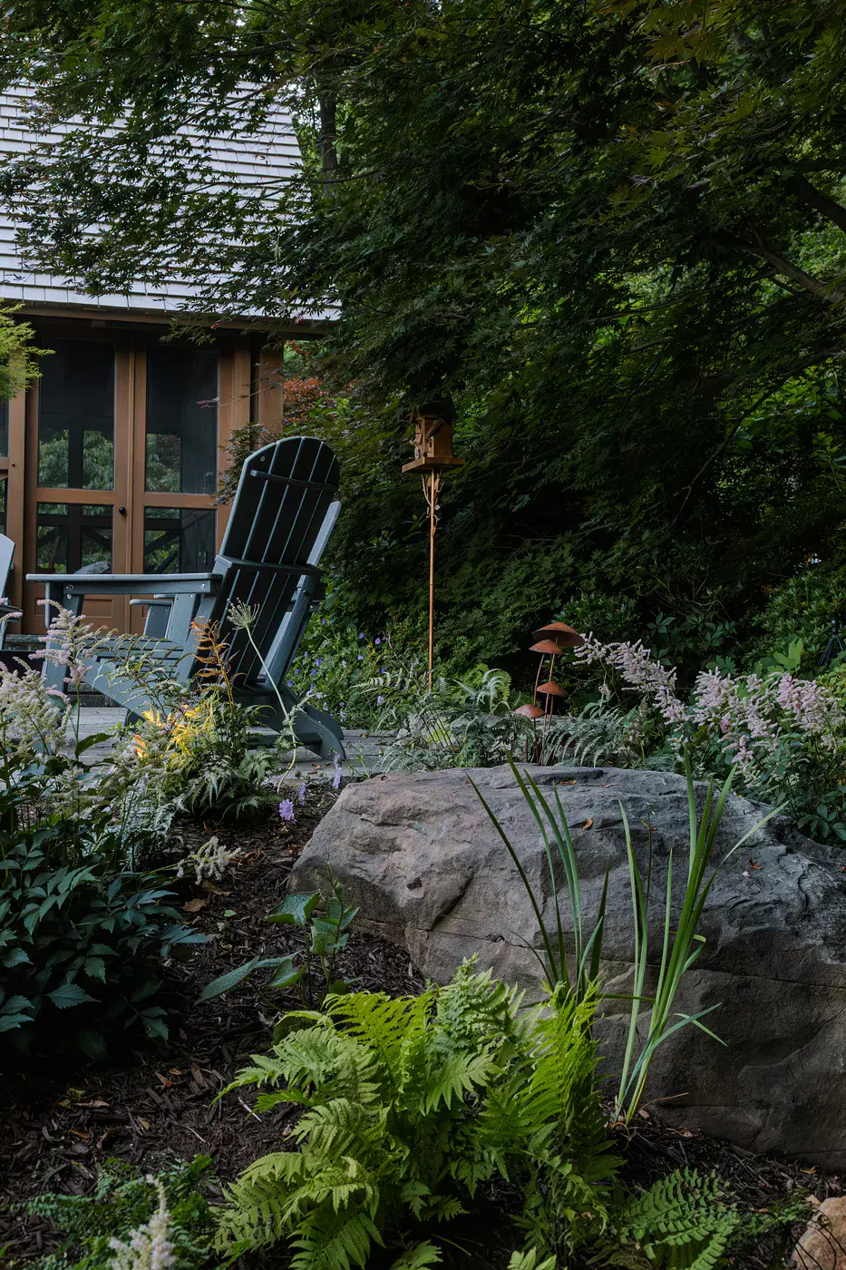 Large natural boulder set among ferns, astilbe, and woodland plantings in a Lincoln University PA garden design, with a gray Adirondack chair, a rusted mushroom sculpture, a birdhouse finial, and the timber-frame screen room beneath a laceleaf maple
